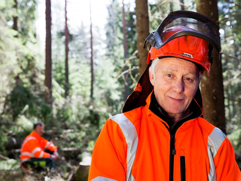 En äldre man i orange skyddskläder och hjälm står i en solig skog, en annan person syns i bakgrunden.