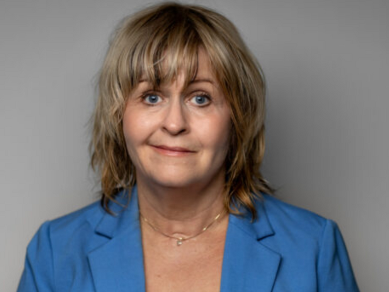 Person med axellångt blont hår, blå kavaj och vit topp mot grå bakgrund.