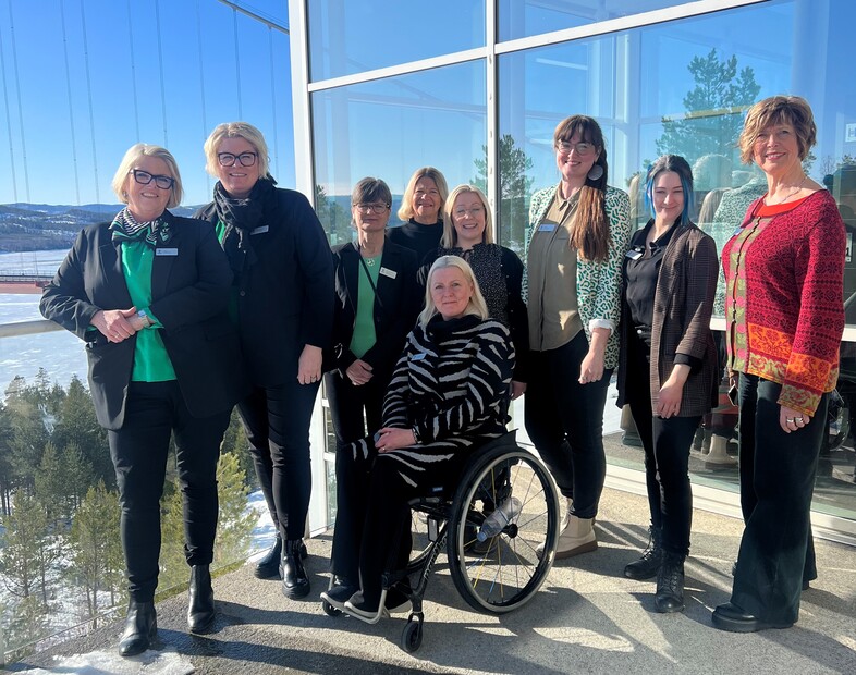 En grupp kvinnor, varav en i rullstol, poserar tillsammans inomhus vid stora fönster med vinterlandskap utanför.