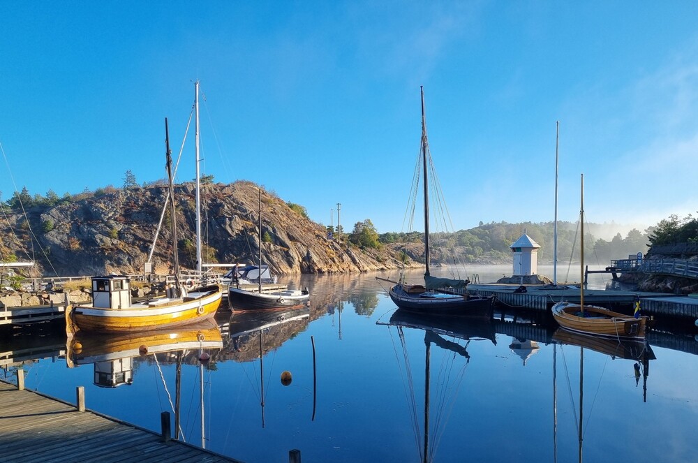Små segelbåtar ligger förtöjda vid en brygga i en lugn hamn med klippor och blå himmel i bakgrunden.