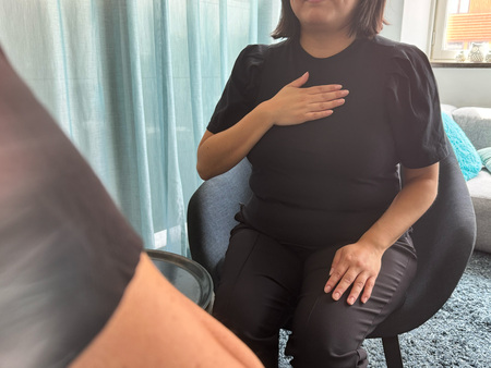 Person sitter på stol med ena handen på bröstet och den andra på låret, inomhus vid ett fönster.