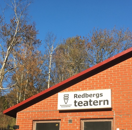 Röd tegelbyggnad med skylt 'Redbergs teatern' mot blå himmel och träd i bakgrunden.