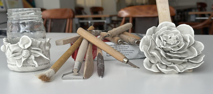 Keramikverktyg, en glasburk dekorerad med lera och en lerblomma på ett bord i ett klassrum.