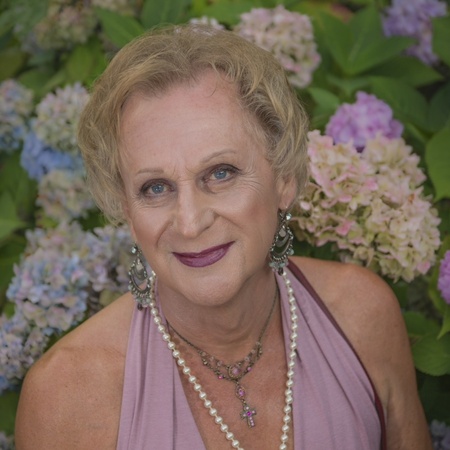 Äldre kvinna med kort blont hår, lila klänning och pärlhalsband poserar framför blommande hortensior.