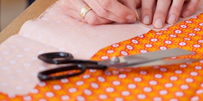 Händer som nålar fast mönsterpapper på orange tyg med sax och krita bredvid.