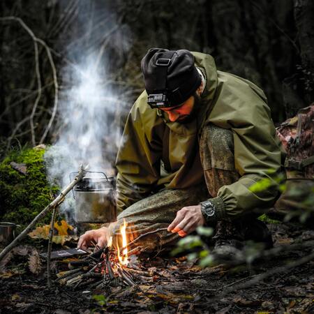 En person i friluftskläder tänder en eld i skogen med en vattenkokare hängande över elden.