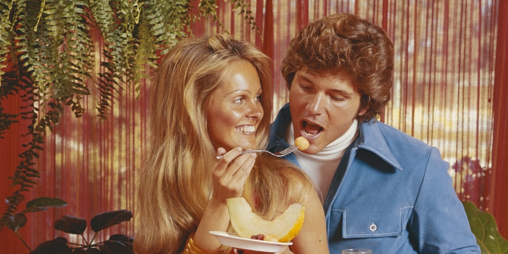 En man och en kvinna står vid ett bord fullt av frukt, mannen håller ett glas juice och båda ler.