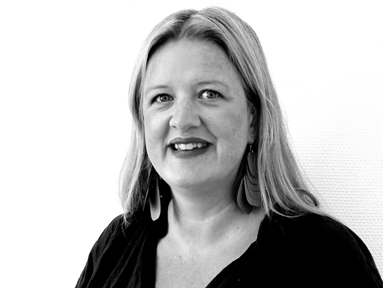 Kvinna med ljust hår och mörk tröja ler mot kameran, svartvit porträttbild mot vit bakgrund.