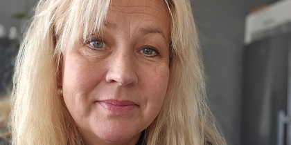 Person med långt blont hår och svart hoodie med orange text i inomhusmiljö.