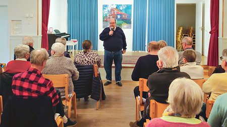 En man står och talar inför en grupp äldre personer i en samlingssal med trästolar och blå gardiner.