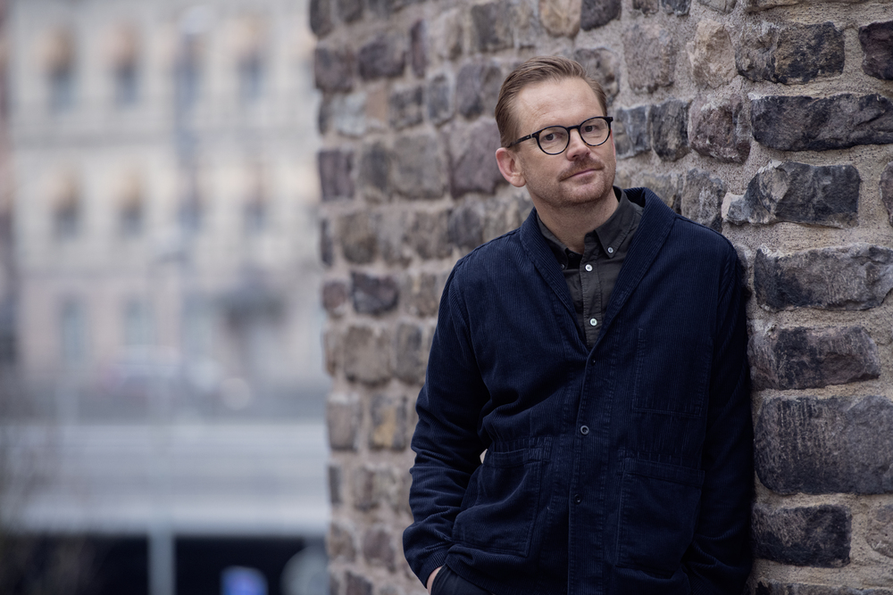 Man med glasögon lutar sig mot en stenmur utomhus, bakgrund med suddiga byggnader.