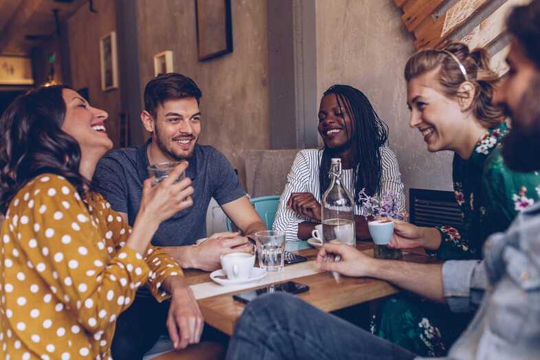 Fem skrattande personer sitter runt ett bord på ett kafé och fikar.