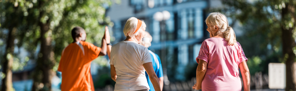 Fyra äldre personer promenerar utomhus i en park med träd och byggnader i bakgrunden.
