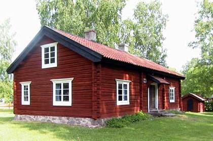 Röd trähus med vita fönster och tegeltak, omgiven av gräs och träd i dagsljus.