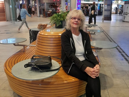 Kvinna sitter på en rund träbänk med små bord i en modern inomhusgalleria med butiker och skyltar.