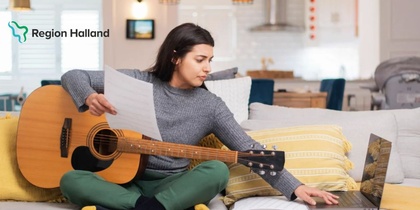 Kvinna sitter i soffa med gitarr, notblad och laptop i ett ljust vardagsrum.