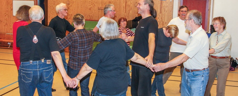 En grupp vuxna håller varandra i händerna och dansar i en ring i en gympasal.