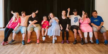 En grupp unga personer sitter på en scenkant och poserar glatt tillsammans inomhus.