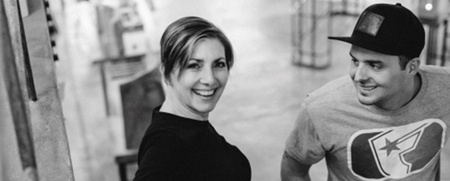 Två personer går uppför en trappa inomhus, en kvinna i svart klänning och en man i t-shirt och keps. Bilden är svartvit.