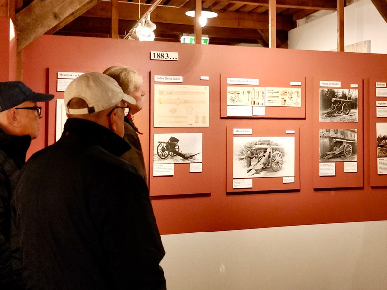 Fem personer tittar på historiska utställningstavlor i ett museum med träbjälkar och rödbruna väggar.