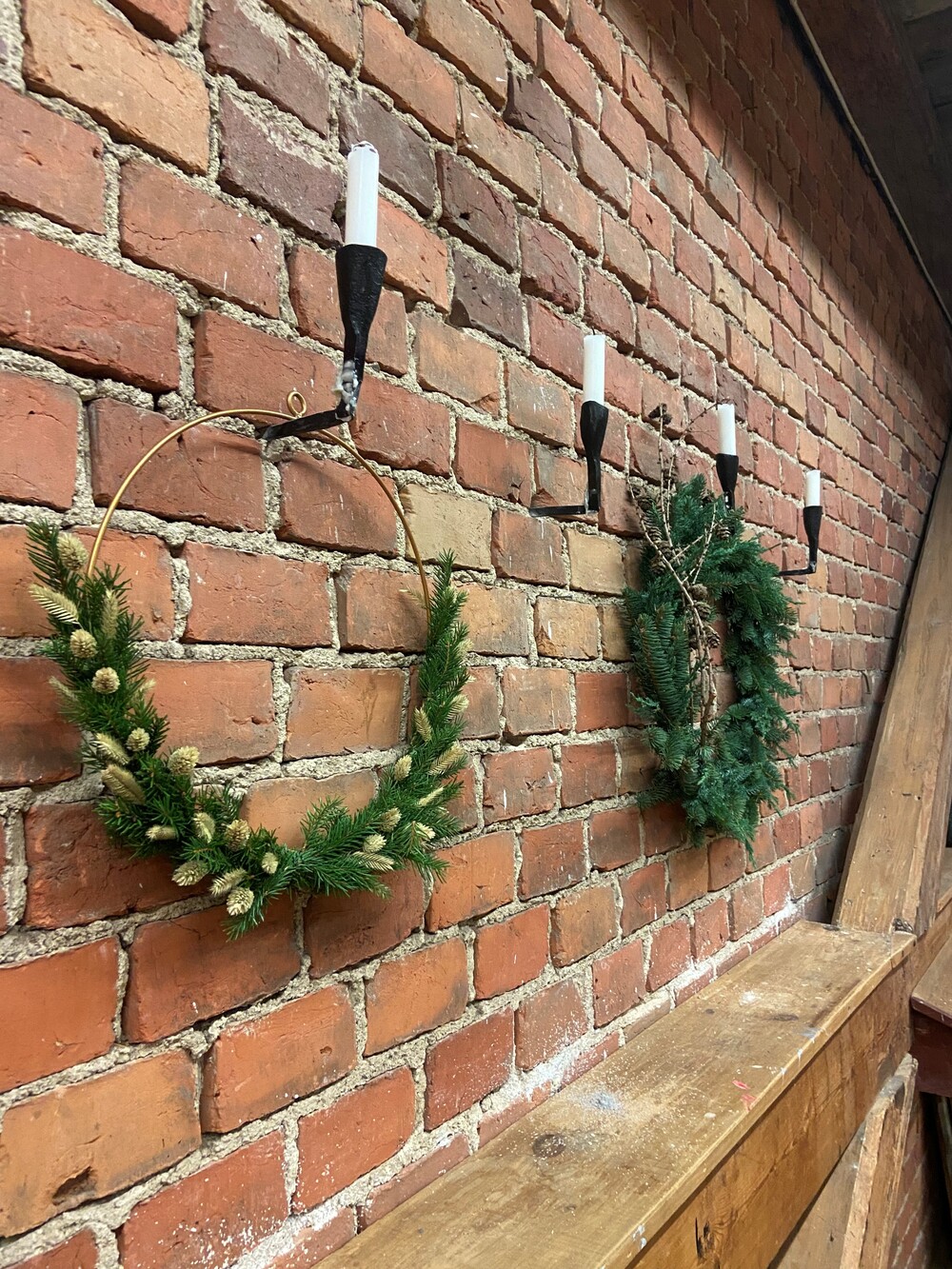 Två julkransar och fyra väggljusstakar med vita ljus på en röd tegelvägg ovanför en träbänk.