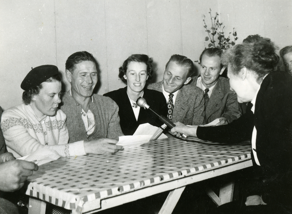 En grupp människor sitter vid ett bord och intervjuas med en mikrofon i en inomhusmiljö.