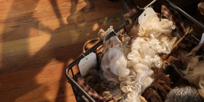 En korg fylld med olika sorters ull i naturfärger, placerad på ett trägolv med lappar fästa på ullen.