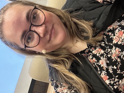 Person med långt ljust hår, blommig topp och svart väst sitter i en stol inomhus.