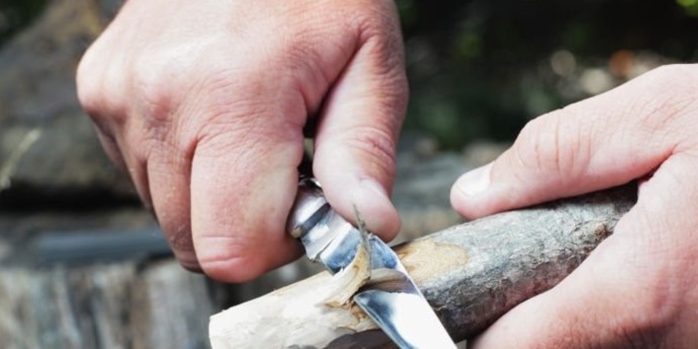 Två händer täljer en träpinne med en fällkniv utomhus.
