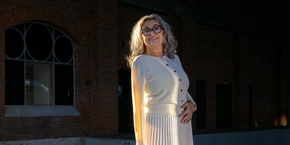 Kvinna i ljus kjol och tröja står på en perrong med tegelvägg och stora fönster i bakgrunden.