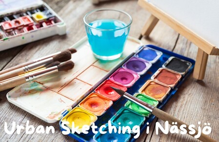 Akvarellfärger, penslar och ett glas vatten på ett träbord med texten Urban Sketching i Nässjö.