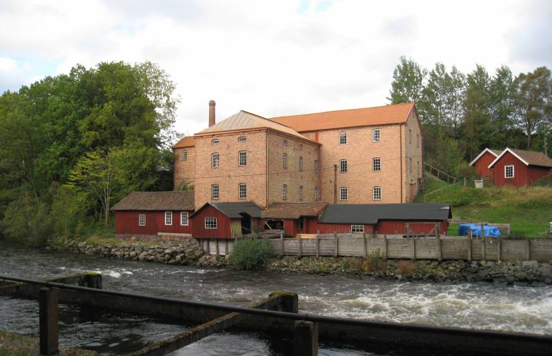 En gammal tegelbyggnad och röda trähus vid en forsande å, omgiven av grönska och träd.