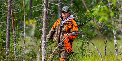 En jägare i orange och kamouflagekläder går med gevär genom en grön skog.