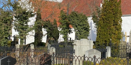 Vit kyrka med rött tak och gravstenar omgiven av järnstaket under blå himmel.
