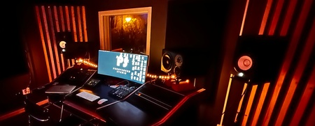 Ett mörkt inspelningsstudio med orange belysning, dator, högtalare och ljudutrustning på ett skrivbord.