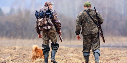 Två jägare i kamouflagekläder och en hund går på ett fält med gevär på ryggen, ryggen mot kameran.