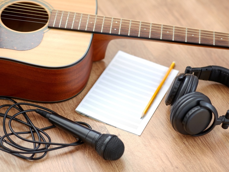 Akustisk gitarr, mikrofon, hörlurar, notpapper och penna på ett trägolv, redo för låtskrivning.