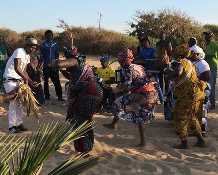 En grupp människor dansar och spelar musik på en sandig plats utomhus, klädda i färgglada traditionella kläder.