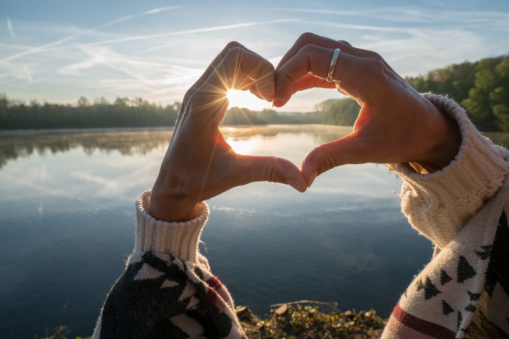 Två händer formar ett hjärta mot en sjö i soluppgång, med skog i bakgrunden.