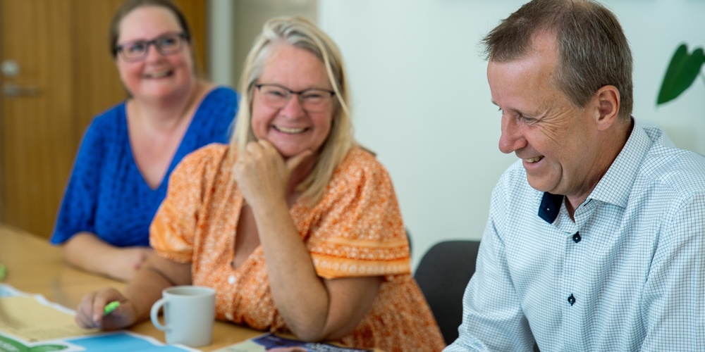 Tre glada vuxna sitter vid ett bord och diskuterar, med papper och pennor framför sig.