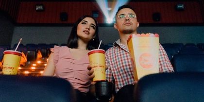 Två personer sitter i en biosalong med popcorn och läsk, filmprojektor lyser bakom dem.