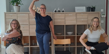 Tre kvinnor poserar lekfullt på och bredvid stolar i ett ljust kontorsrum med hyllor i bakgrunden.