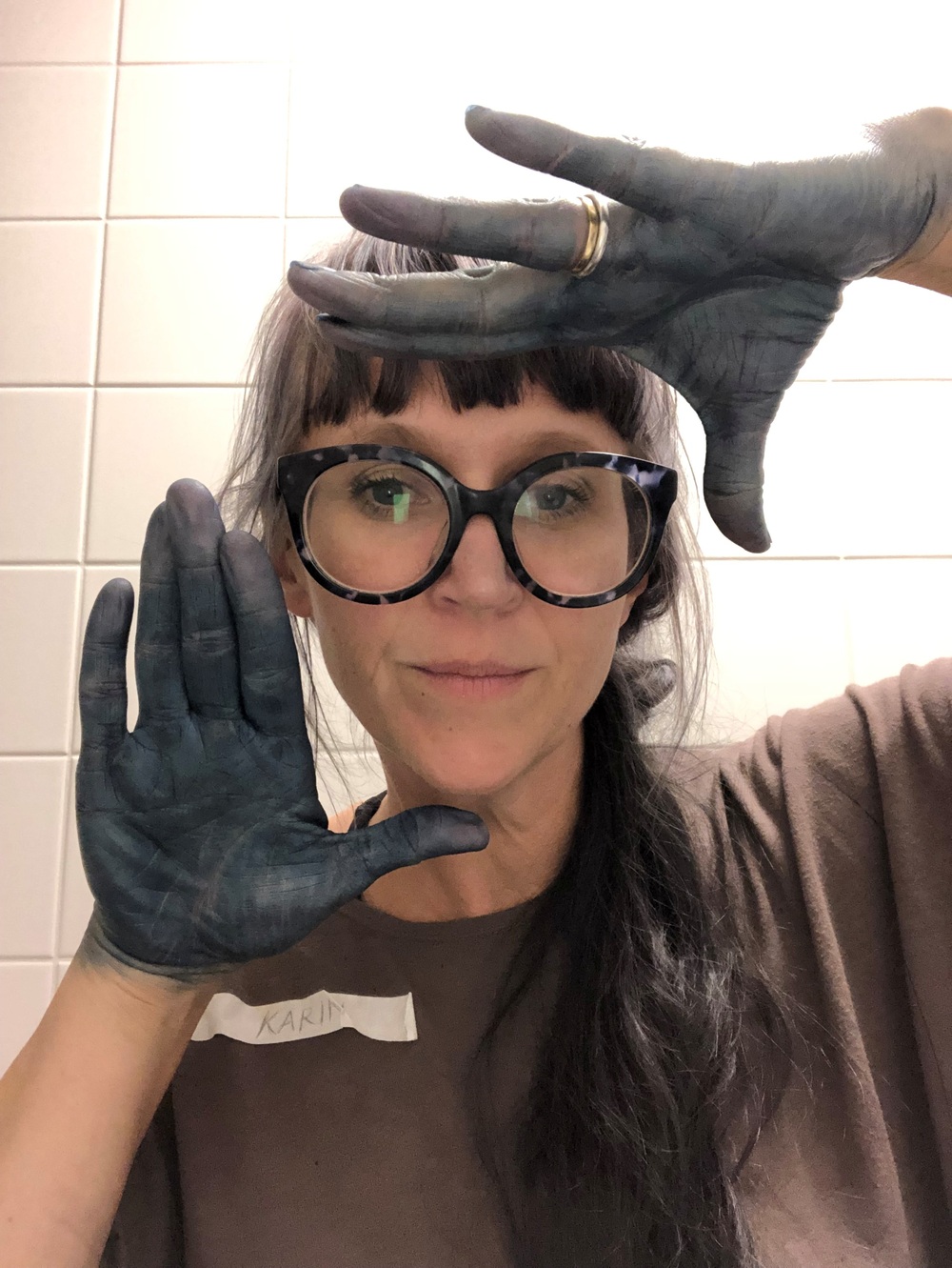 Kvinna med stora glasögon visar sina blåfärgade händer framför ansiktet, står mot vit kakelvägg.