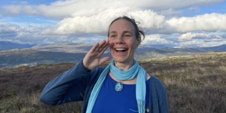 Person står utomhus på fjället med blå kläder och halsduk, håller handen vid huvudet som en hälsning.