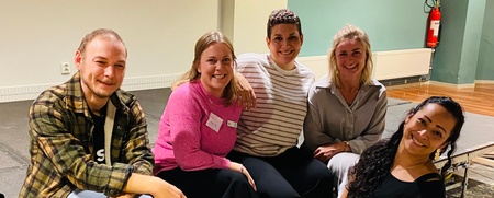 Fem personer sitter tillsammans på en scen och ler mot kameran i en inomhusmiljö.