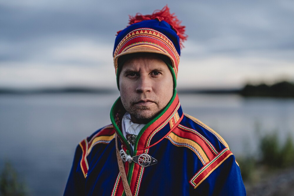 Person i traditionell samisk dräkt står vid en sjö med suddat ansikte.