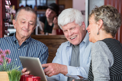 Tre äldre personer sitter vid ett bord på ett café och tittar på en surfplatta tillsammans.