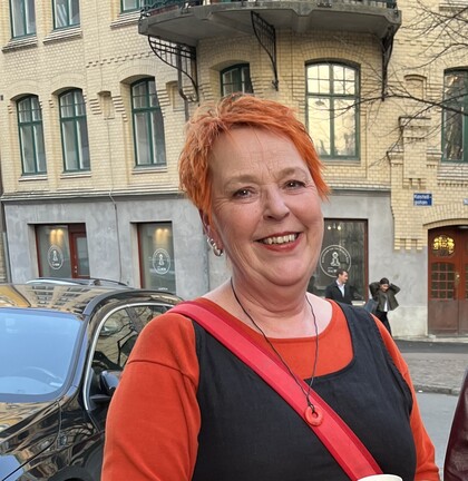 Person med kort orange hår och svart klänning står utomhus framför en gul tegelbyggnad.