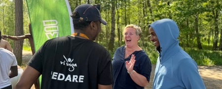 Tre personer samtalar och skrattar utomhus vid en grön flagga i en solig skogsmiljö.