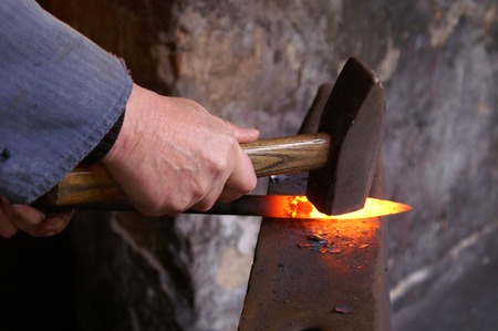 En person smider en glödande metallbit med en hammare på ett städ.